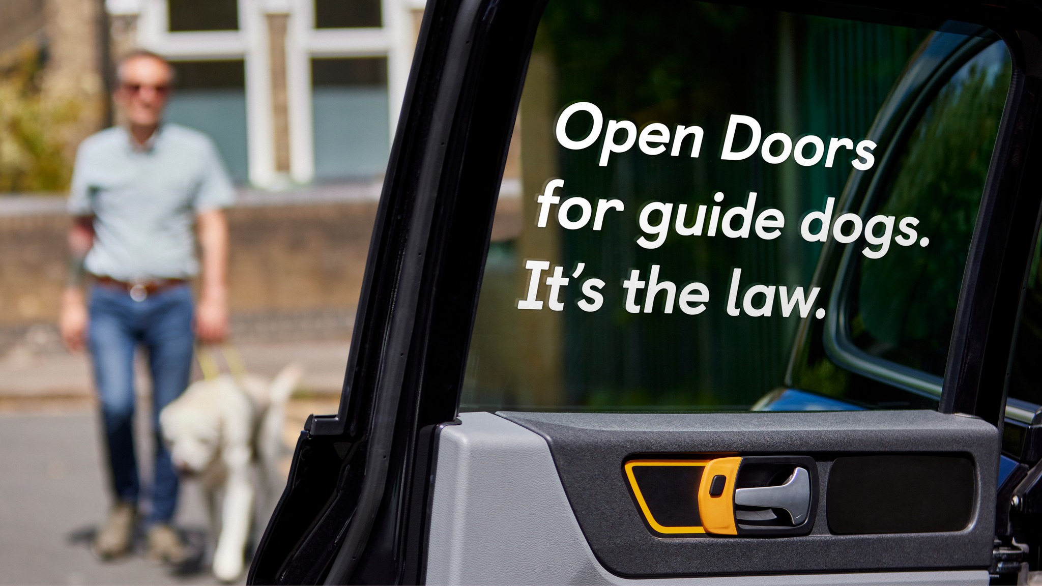 A woman in a blue top hands a sticker to someone. The sticker is yellow and blue and reads, 'Open Doors for guide dogs and all other assistance dogs'.