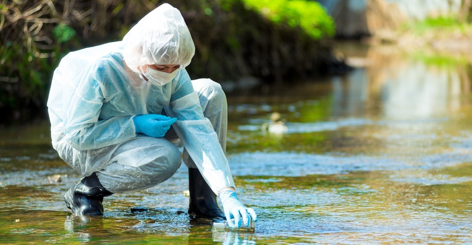 Scientist in protective suit takes water for analysis from polluted river.