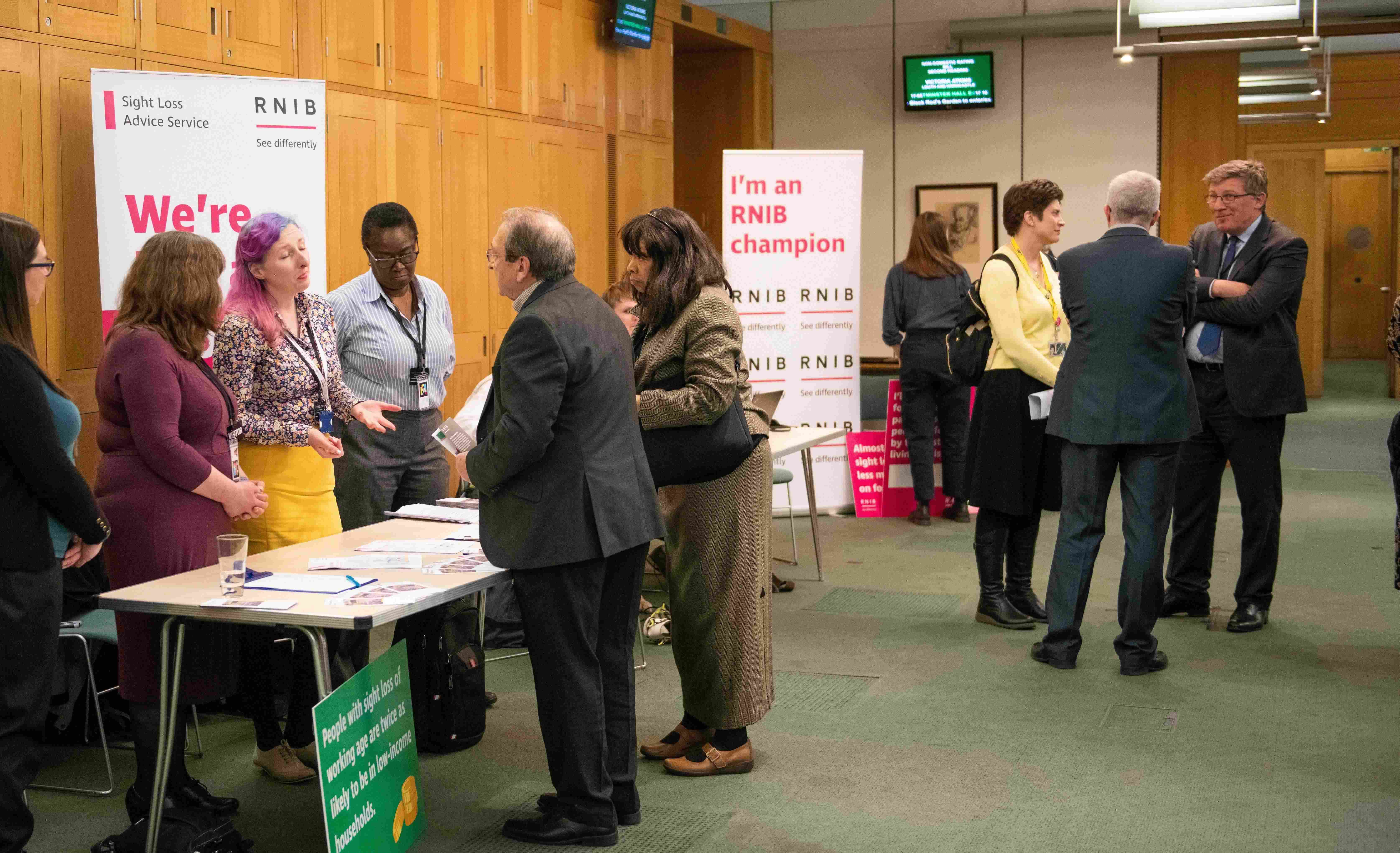 RNIB parliamentary drop-in event where MPs are talking to RNIB staff at across a table. There's a sign in the background which says "I'm an RNIB Champion".