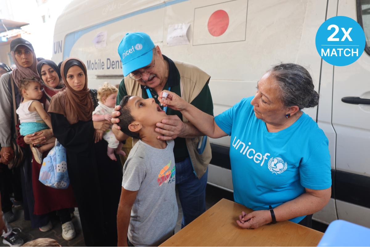 Two UNICEF vaccinators help a child receive an oral dose of the poliovirus vaccine, with a line-up of families waiting in the background.