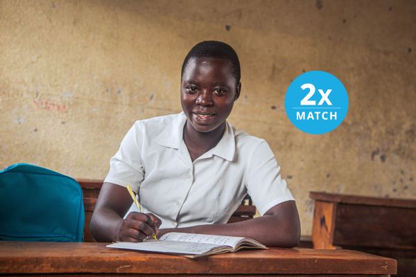 Portrait of a student sitting at a school desk holding a pencil over an exercise book and looking at the camera with a neutral expression, with the overlaid copy 2X Match.