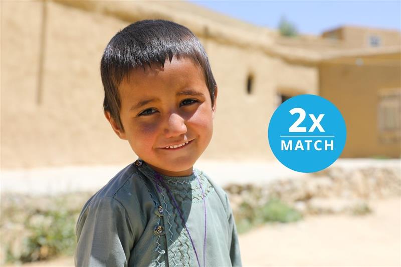 Portrait of a young child smiling at the camera in a sunny outdoor setting in front of a clay building.