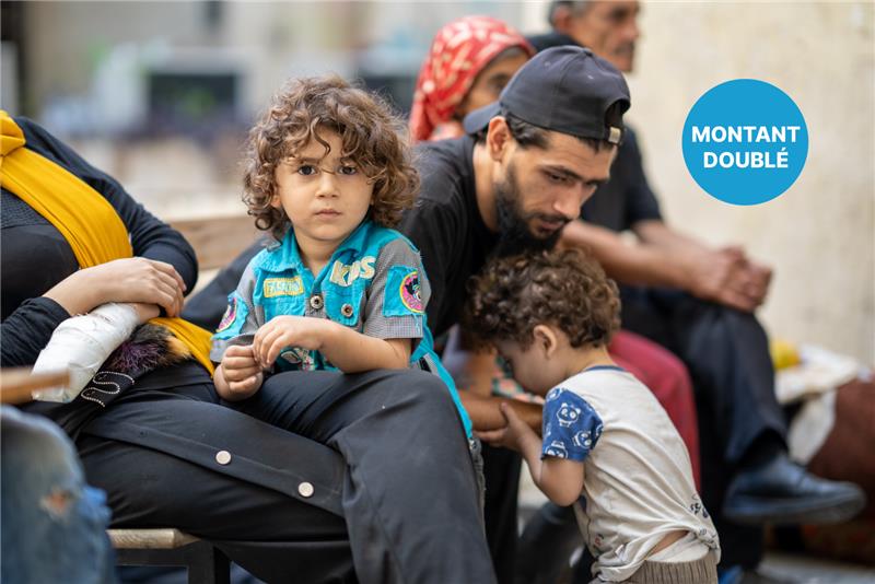 Un enfant regarde l’objectif d’un air neutre, assis sur un banc, en compagnie d’adultes et d’un enfant en bas âge.