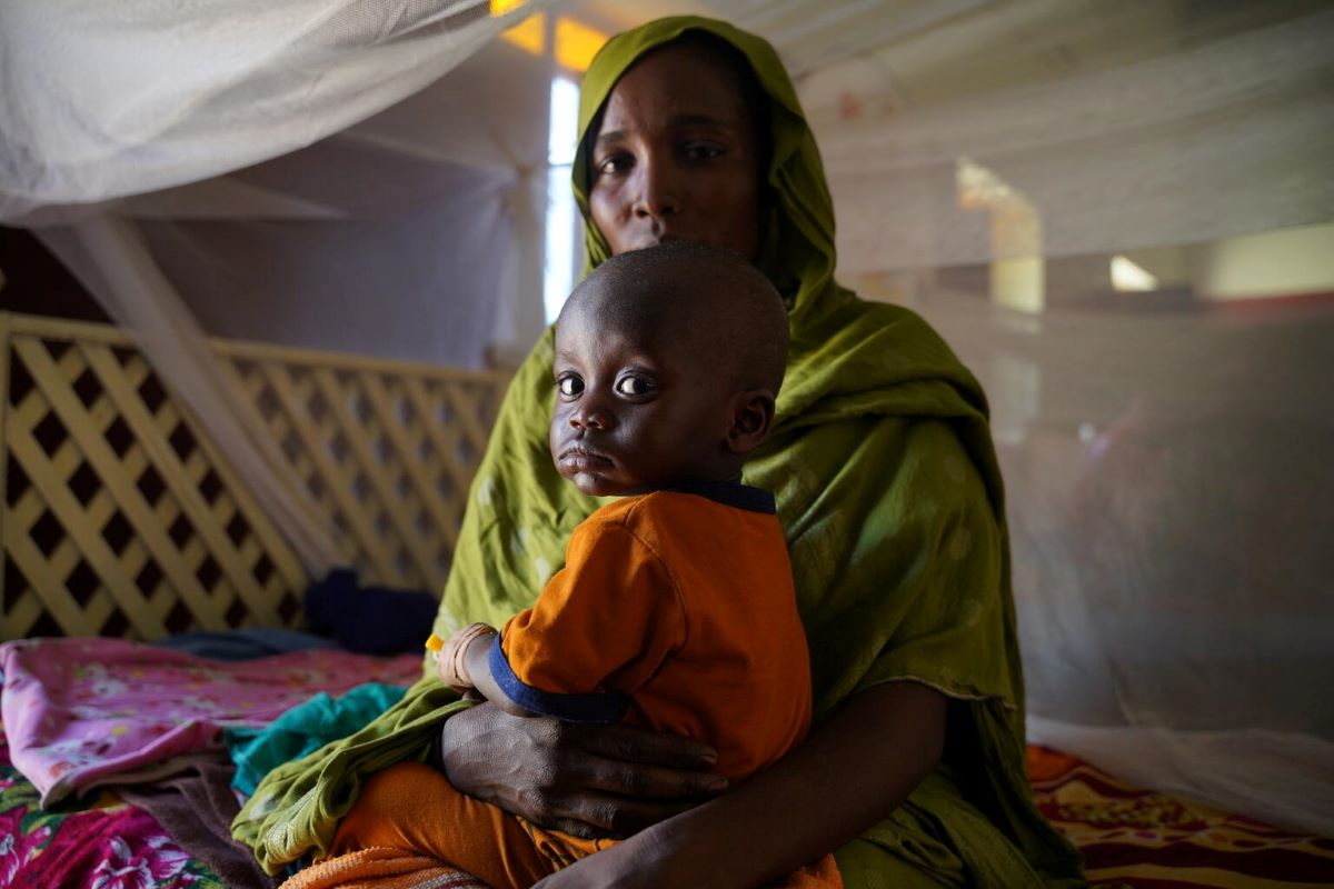 A woman and child sit on a bed in a tent. The child looks directly at the camera with a sad expression.