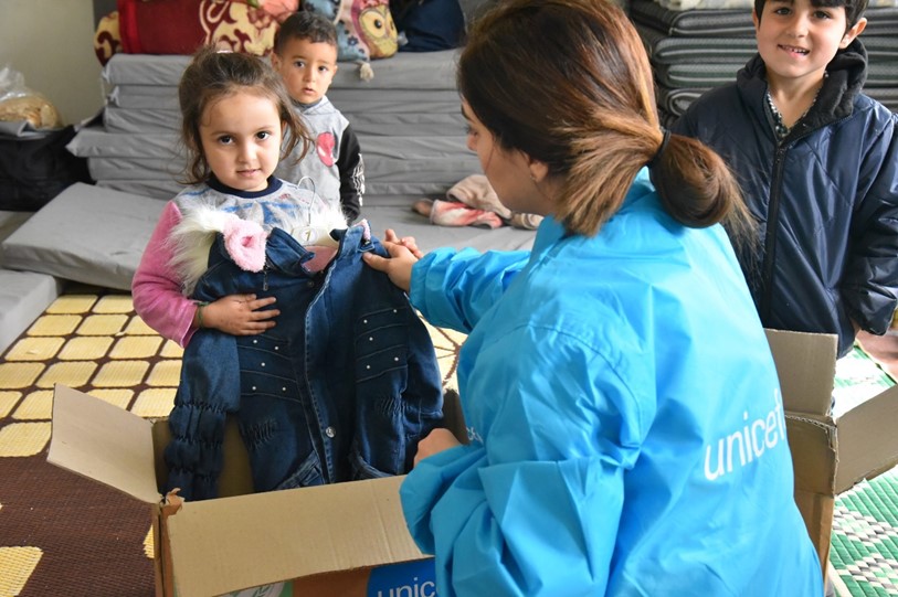 A smiling young girl is presented with a winter jacket by a UNICEF worker while two other children stand behind her.