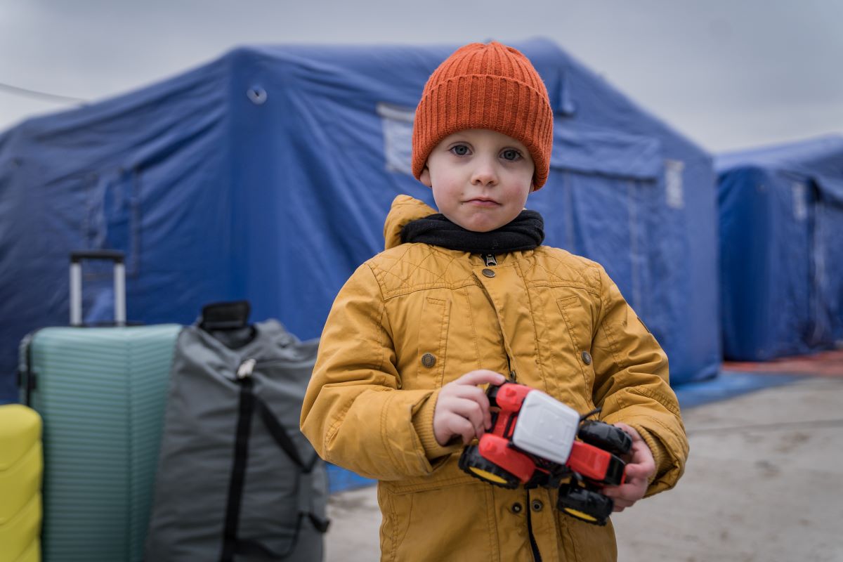 Un garçonnet de quatre ans vêtu de vêtements d’hiver se tient devant des tentes bleues, tenant dans ses mains une voiture-jouet. 
