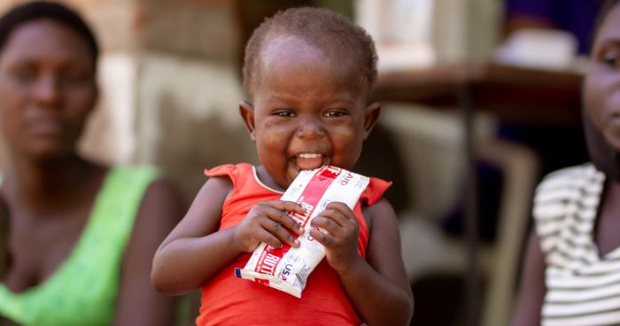 Two-year-old girl smiles widely at the camera while eating Plumpy’Nut at a hospital in Malawi. 