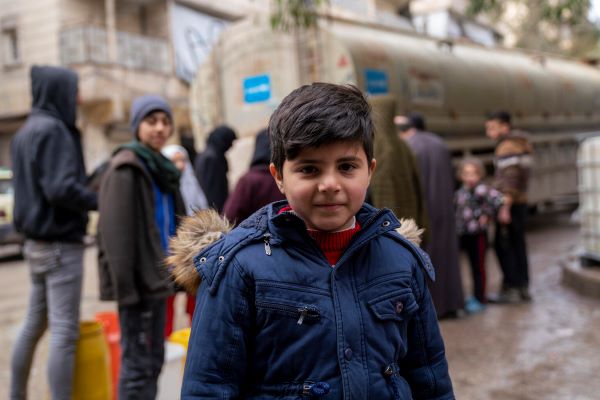 A boy stands in front of a UNICEF water truck, and other people stand behind him in a queue to fill water.