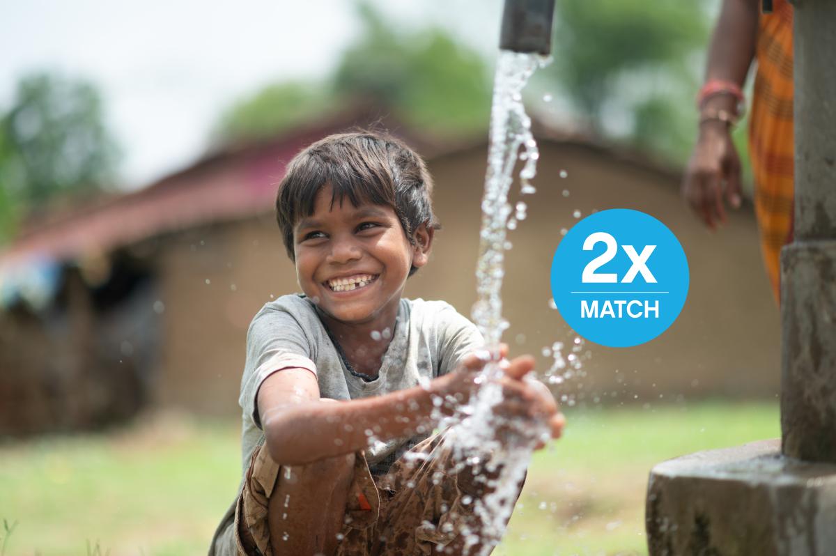 A boy smiles at the camera while washing his hands at a water pump.