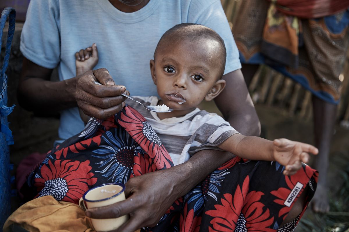 A child in the arms of a care taker being fed while looking towards camera.