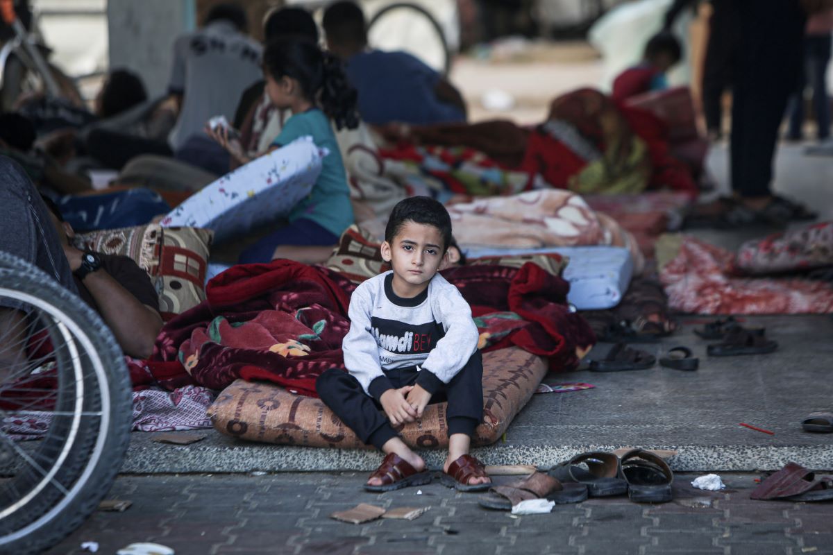 A young boy sitting outside on a mattress looking at the camera with several other displaced children in the background.