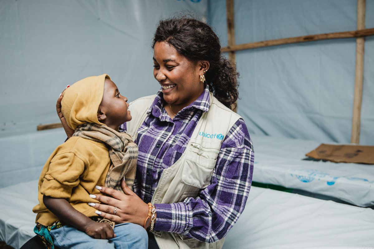 A UNICEF worker holding a child on their lap as they smile at each other while sitting in a tented shelter.