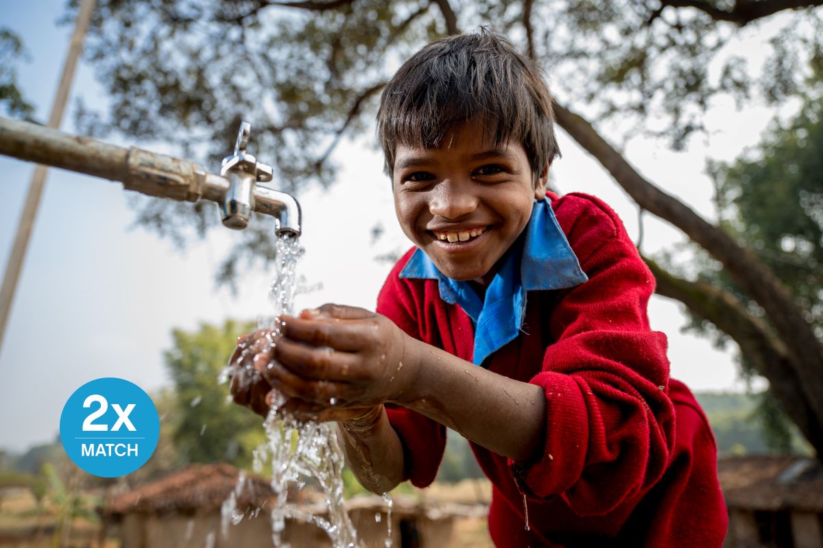 Portrait of a child washing their hands underneath an outdoor waterspout while smiling at the camera.