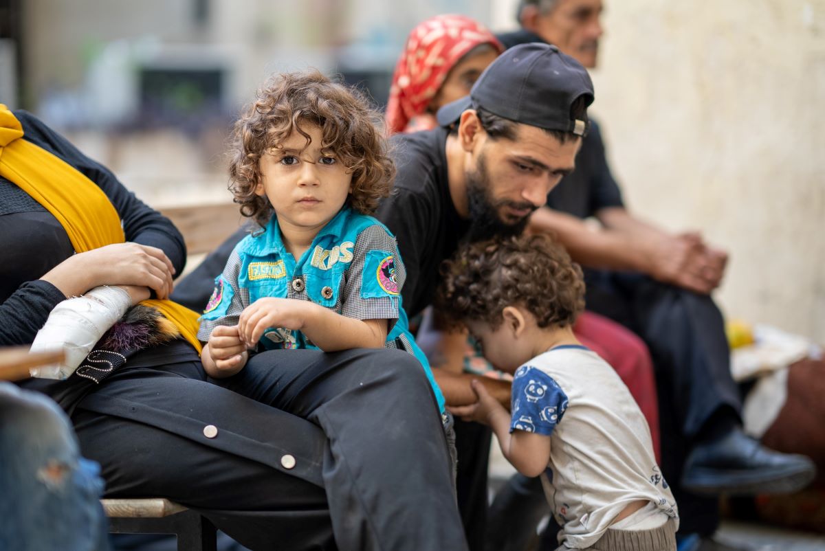 A child looking at the camera with a neutral expression while sitting on a bench next to adults and a toddle.