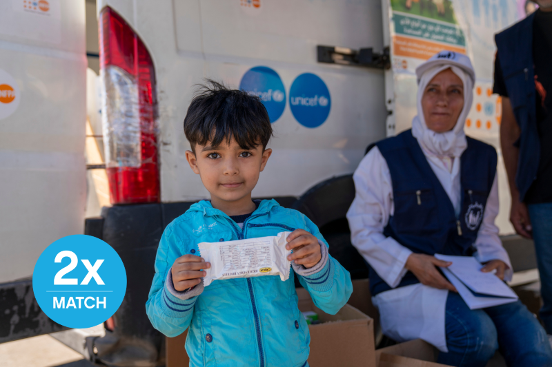 : A child looking directly at the camera with a calm expression while holding up a packet of therapeutic food at a UNICEF centre.