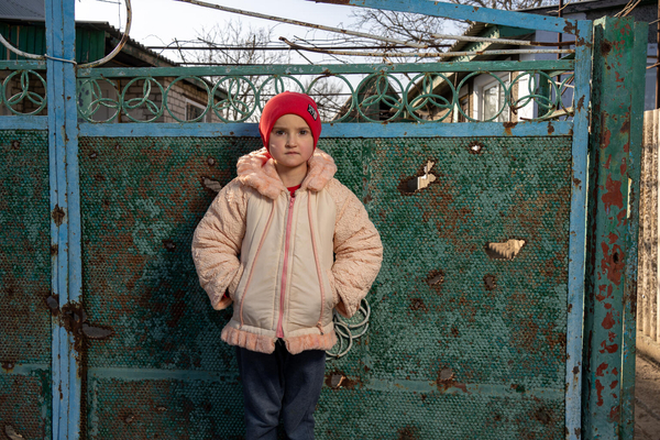 In Ukraine, a girl wearing a red hat stands in front of a metal gate with shrapnel holes.