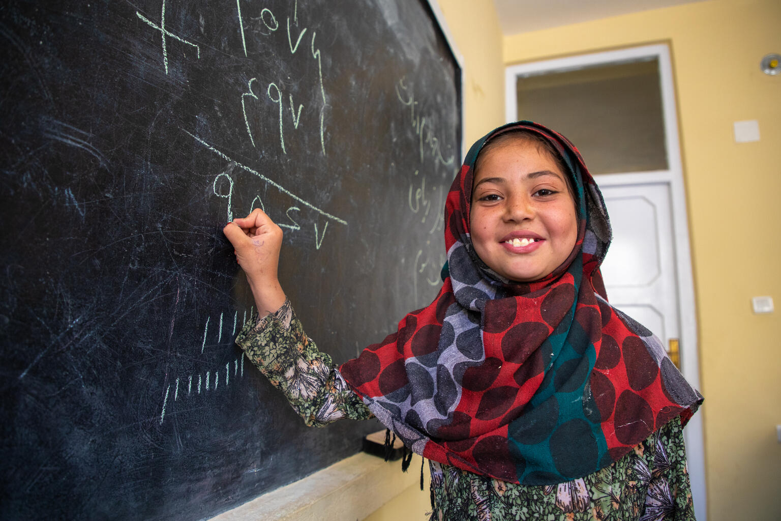 Smiling young girl wearing a headscarf writes math problems on a classroom chalkboard.