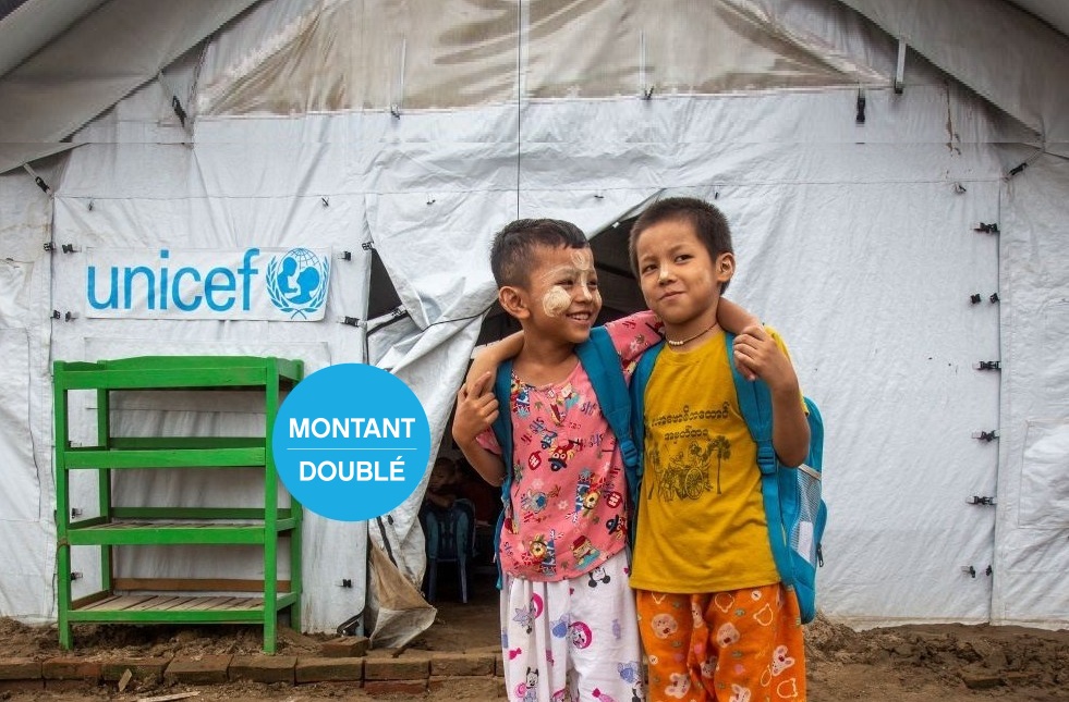 Deux enfants portant des sacs à dos se tiennent devant une tente temporaire de l’UNICEF, souriants, bras dessus bras dessous.