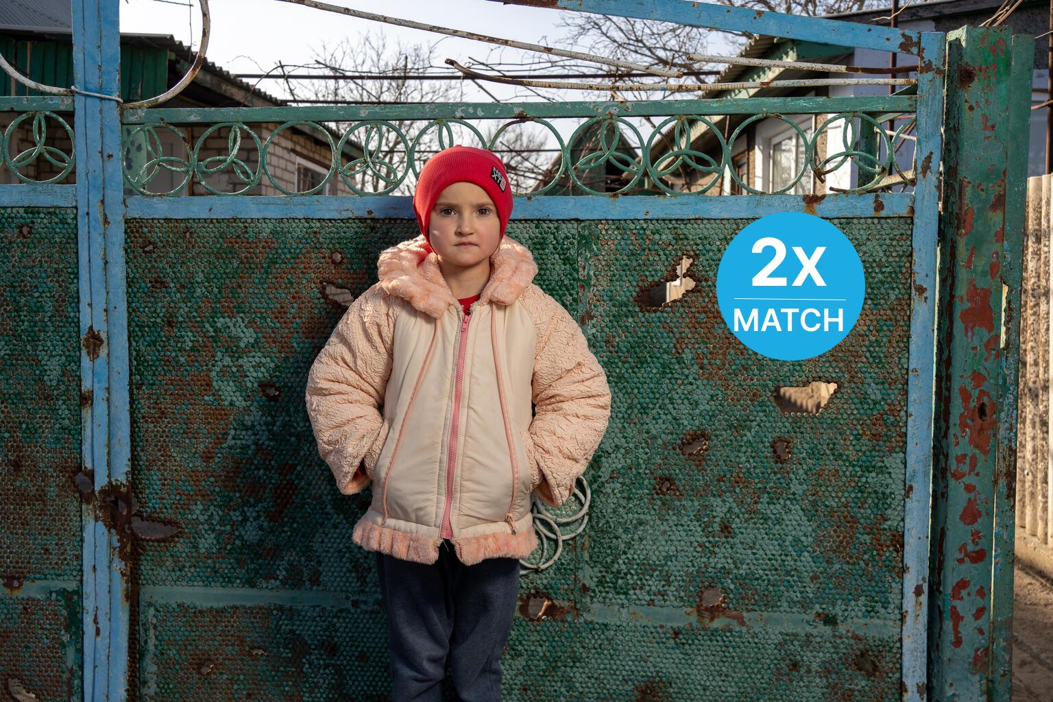 In Ukraine, a girl wearing a red hat stands in front of a metal gate with shrapnel holes.