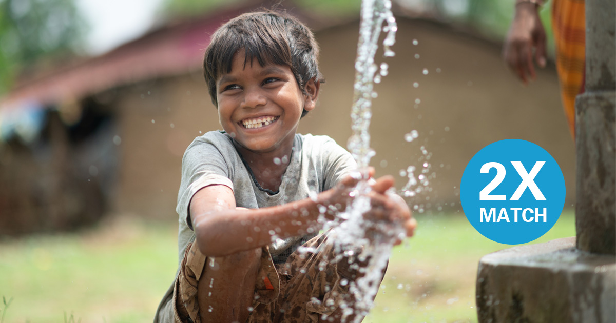 A boy smiles while washing his hands at a water pump.   