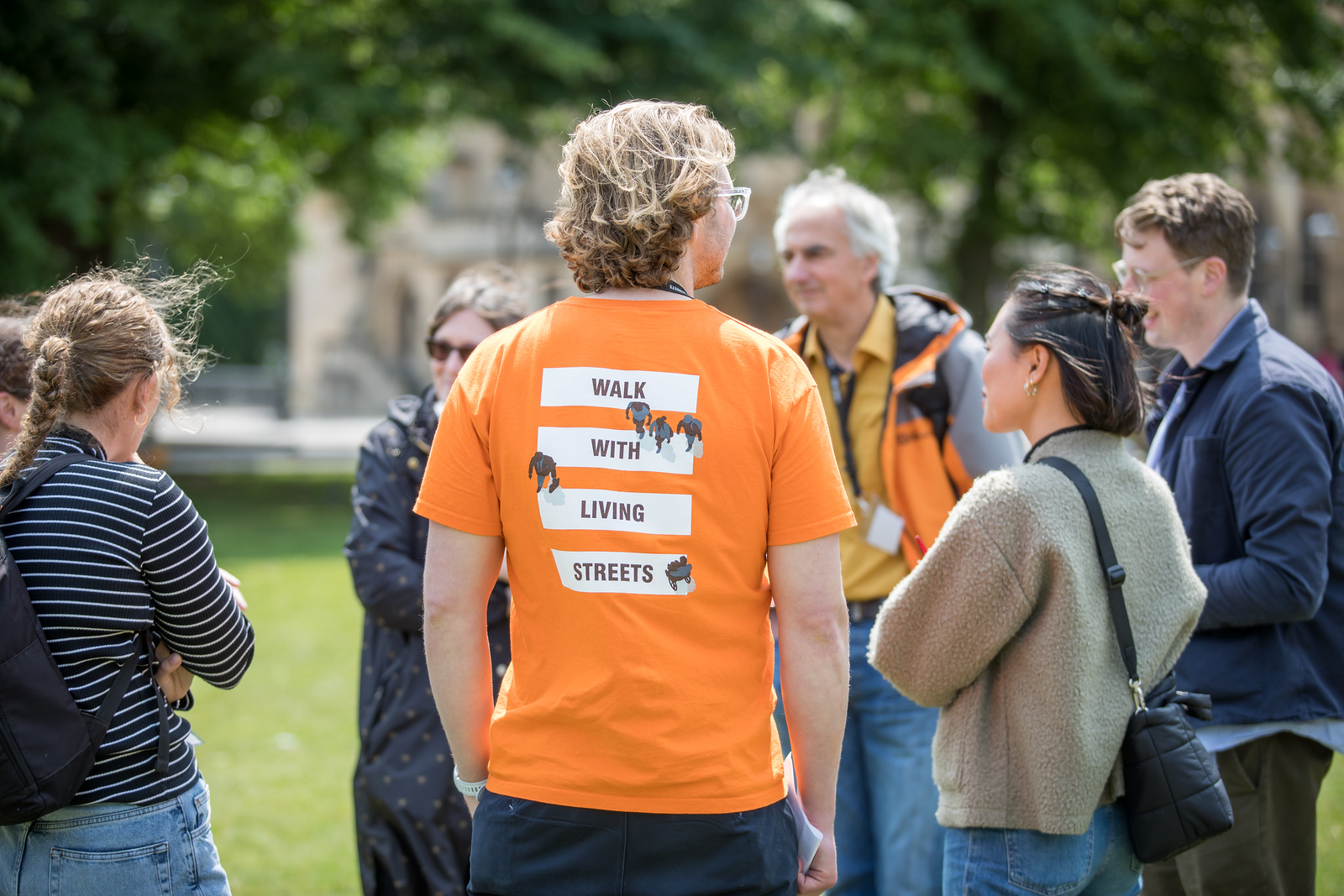 Someone standing with their back to the camera. They are standing as the focal point in a group of people. They are wearing a bright orange tshirt which reads 