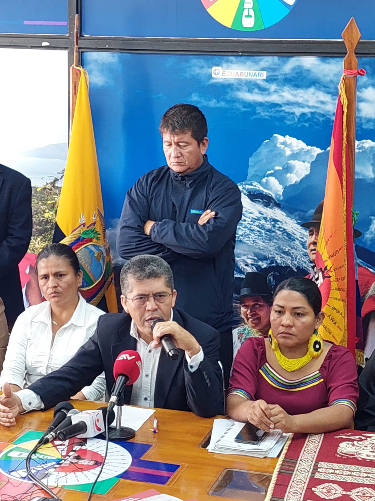 Pablo Fajardo Mendoza is seated and is speaking into a microphone. There are several other microphones in front of him. He is flanked by two women and there is a man standing behind him.