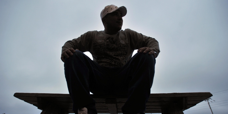 A Hispanic man wearing a baseball cap sits on a picnic table with his hands resting on his knees. His body in silhouette to hide his identity.