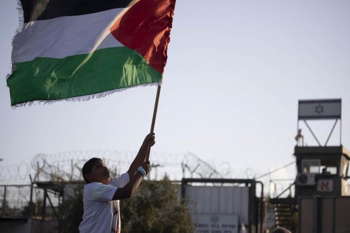 Boy runs with a Palestinian flag in front of a prison with the Israel flag on it.