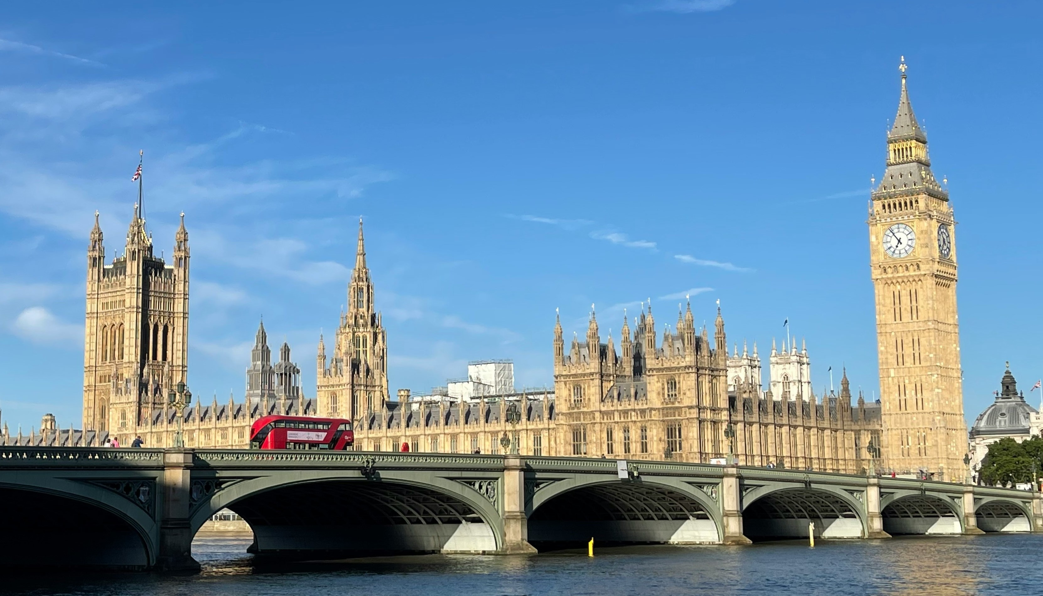 The Palace of Westminster on a sunny day with a blue sky. There is a red double decker bus going over the bridge.