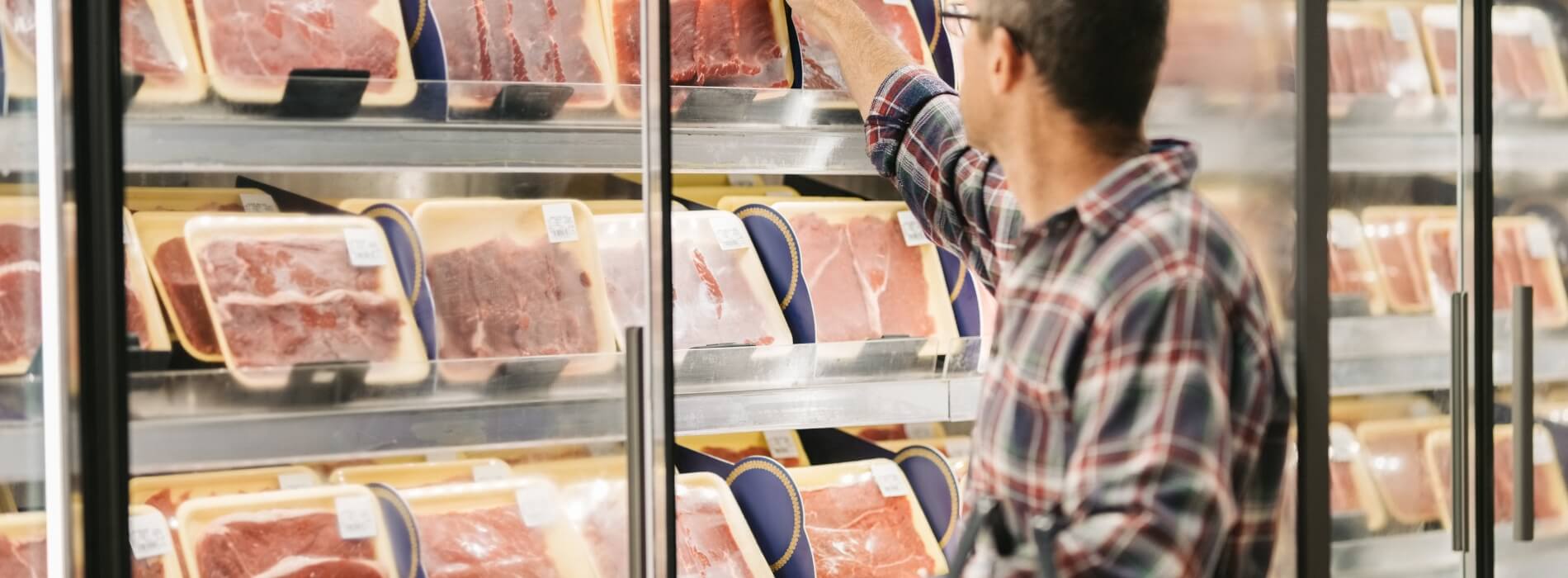 A man reaching into a refrigerator full of meat packages at a grocery store.