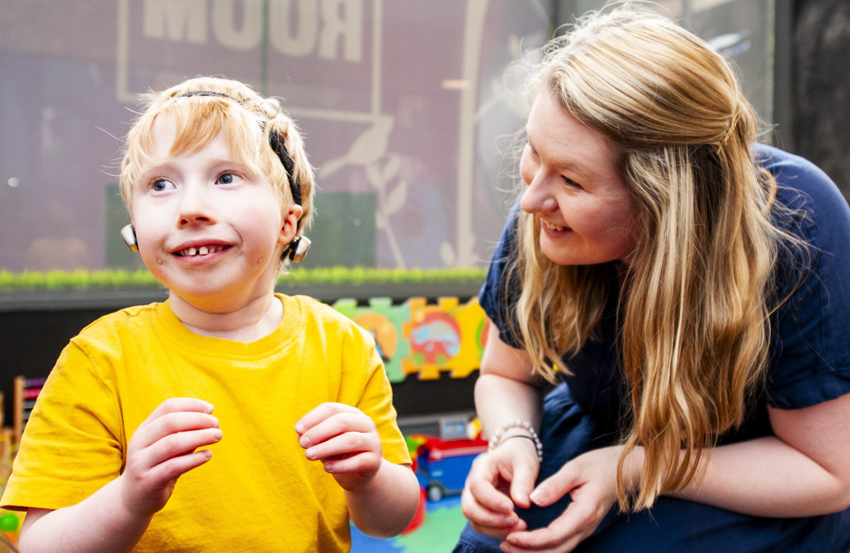 A white woman with long blonde hair sitting next to and looking at a white, blonde boy with microtia who is wearing bone anchored hearing aids on a headband.