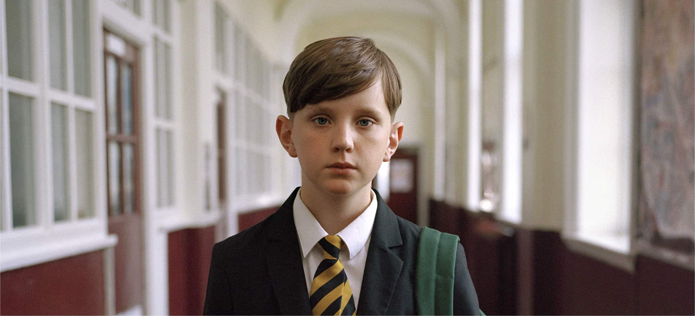 Young boy in school uniform standing in a hallway.