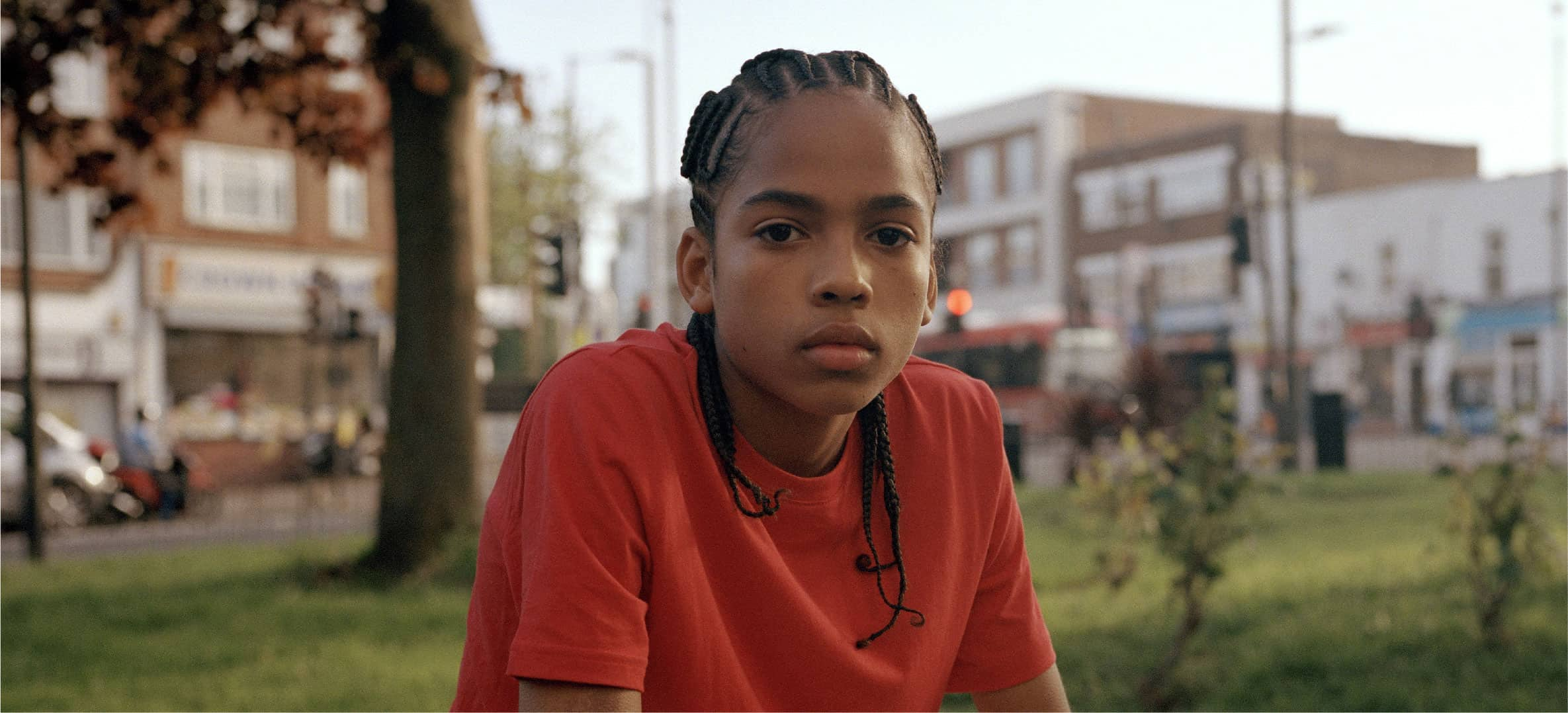 Young boy in a red shirt with braided hair sitting outside in an urban area.




