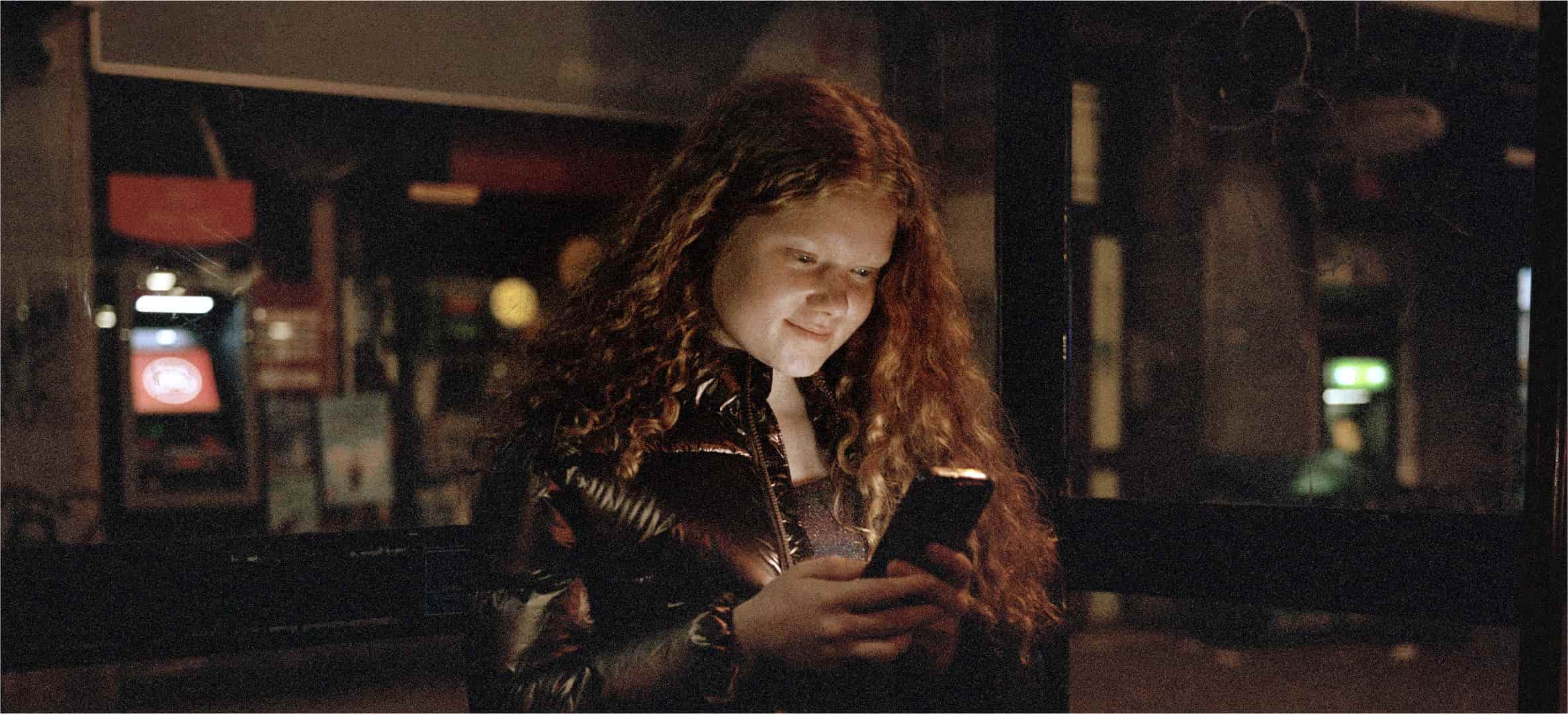 A young woman with curly red hair smiles at her phone at night, wearing a shiny dark jacket.