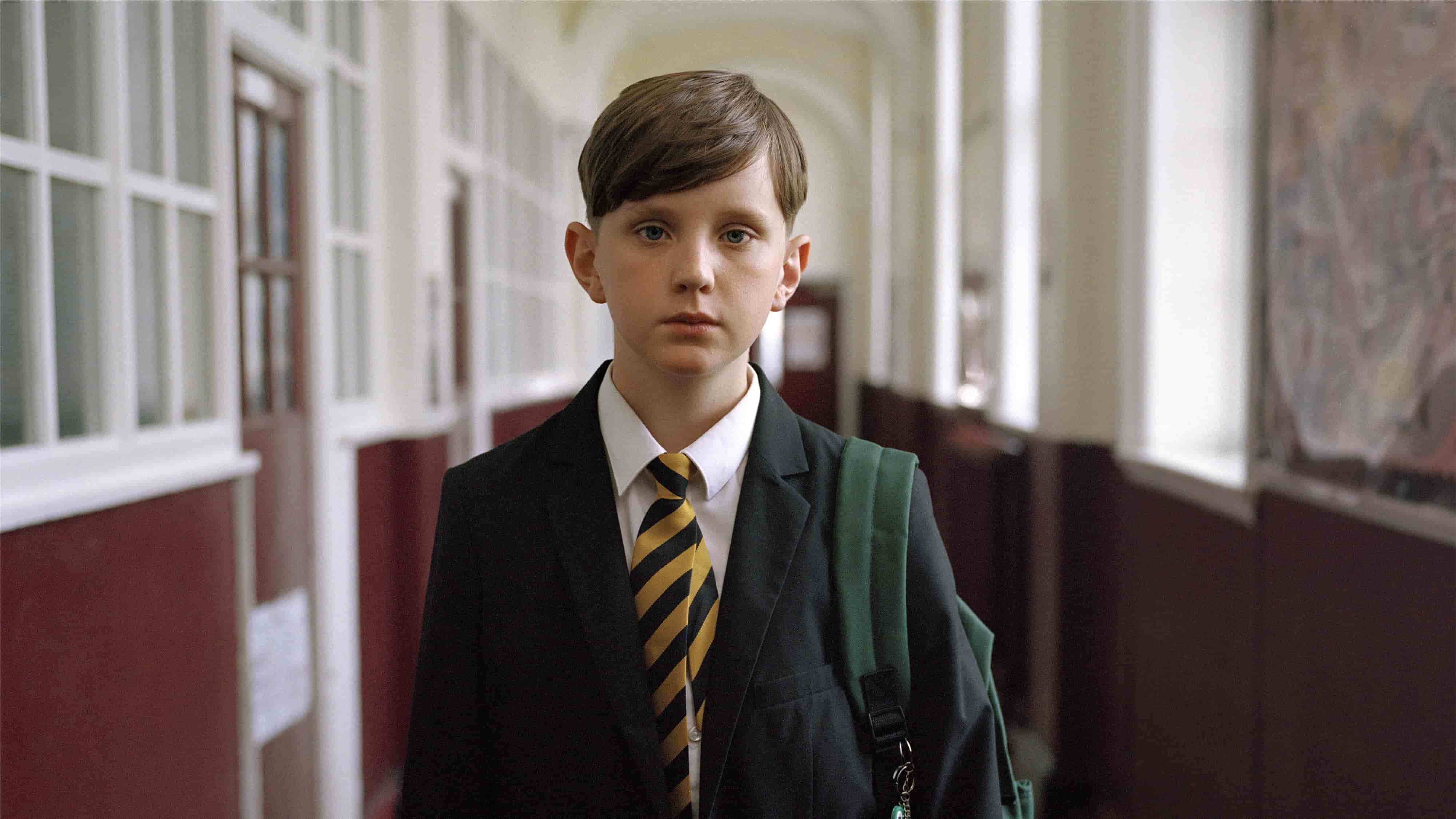A young boy in a school uniform with a striped tie and green backpack.