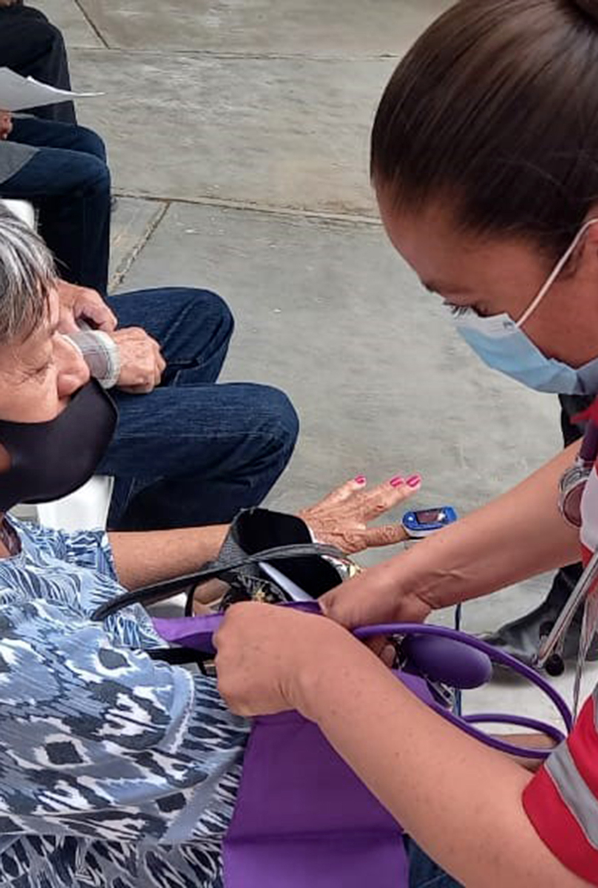A Red Cross worker provides medical care to an elderly woman, both wearing surgical masks.