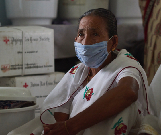 A woman wearing a medical mask sits surrounded by Indian Red Cross Society boxes.