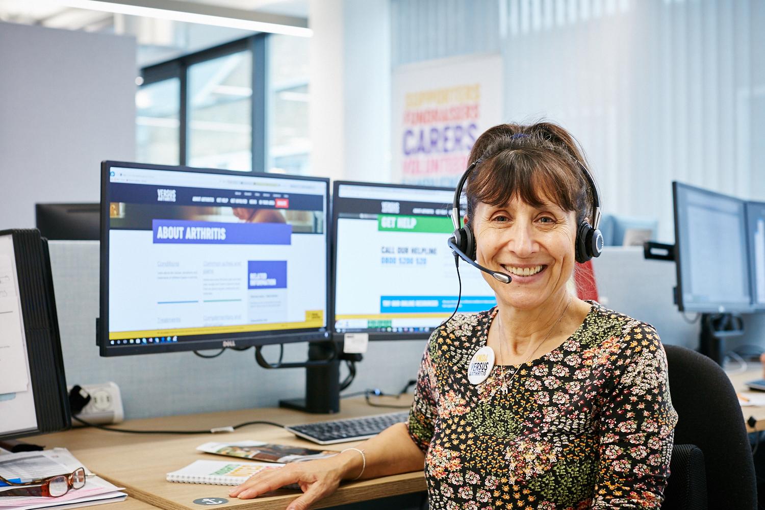 Jayne, a Versus Arthritis Helpline advisor, is sitting at her desk, smiling to the camera. She has a headset on and you can see the Versus Arthritis website on her monitor behind her.