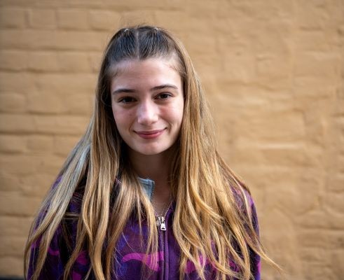 Girl wearing a purple top, standing infront of a cream wall smiling at the camera