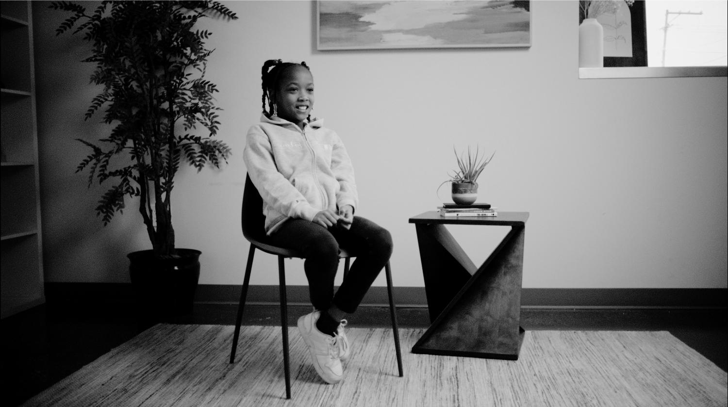Mesiah, an elementary school-aged Black girl, sits in a chair and looks off camera to the right.