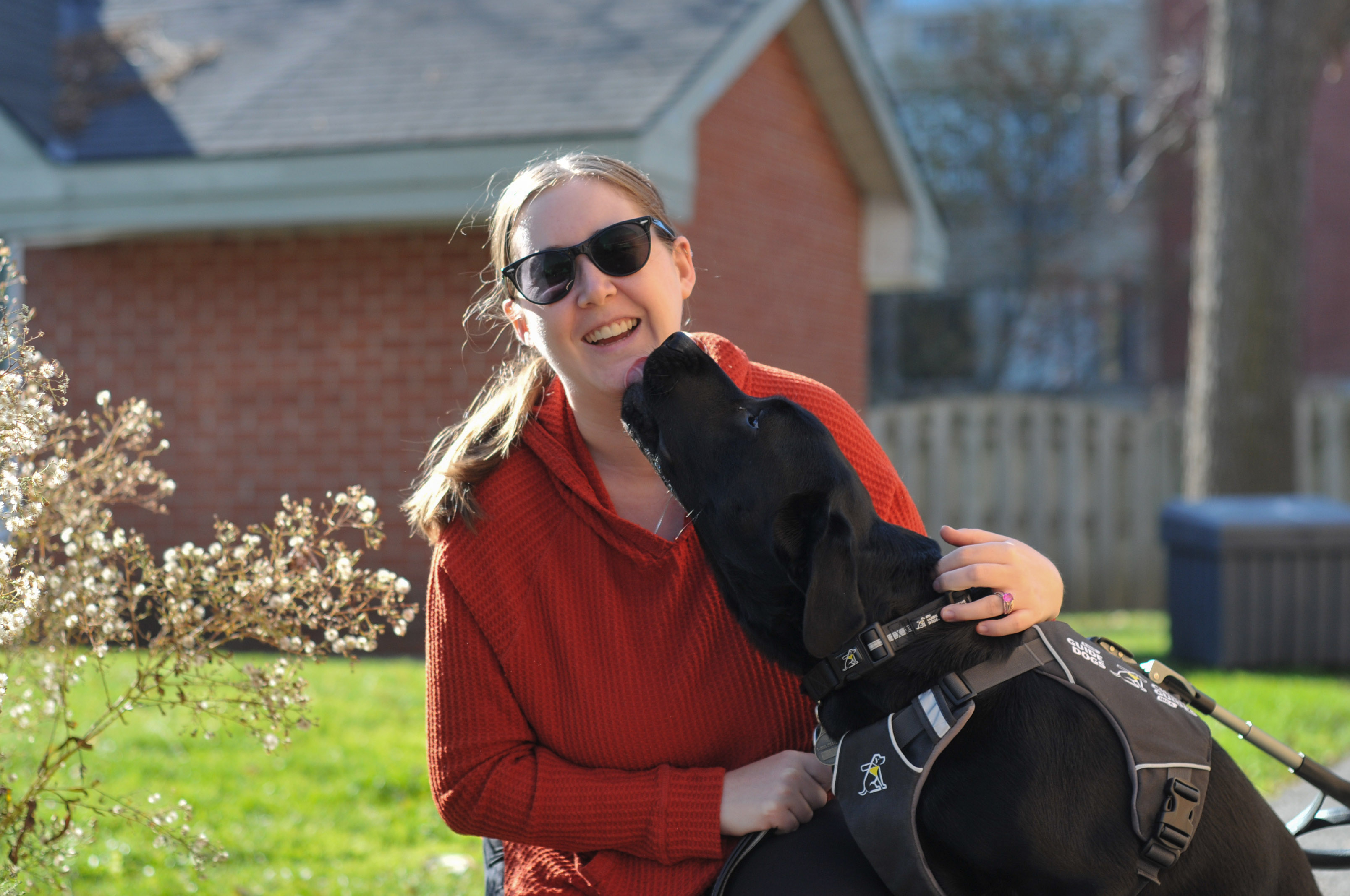 Christine kneeling with her CNIB Guide Dog, Edie. Edie is licking Christine's face who is smiling at the camera.