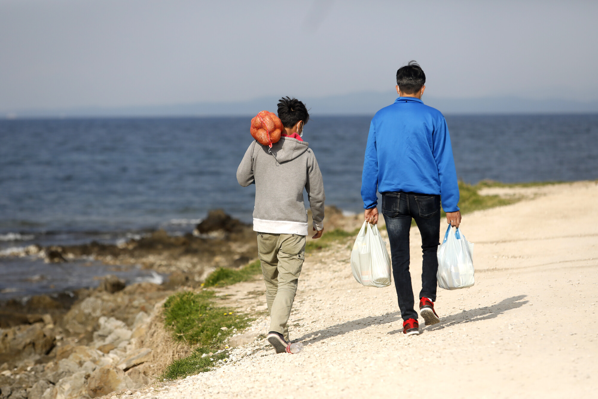 A man and a boy carrying bags of grocery, next to a body of water, and walking to their destination, in the largest refugee camp in Europe, in Moria.