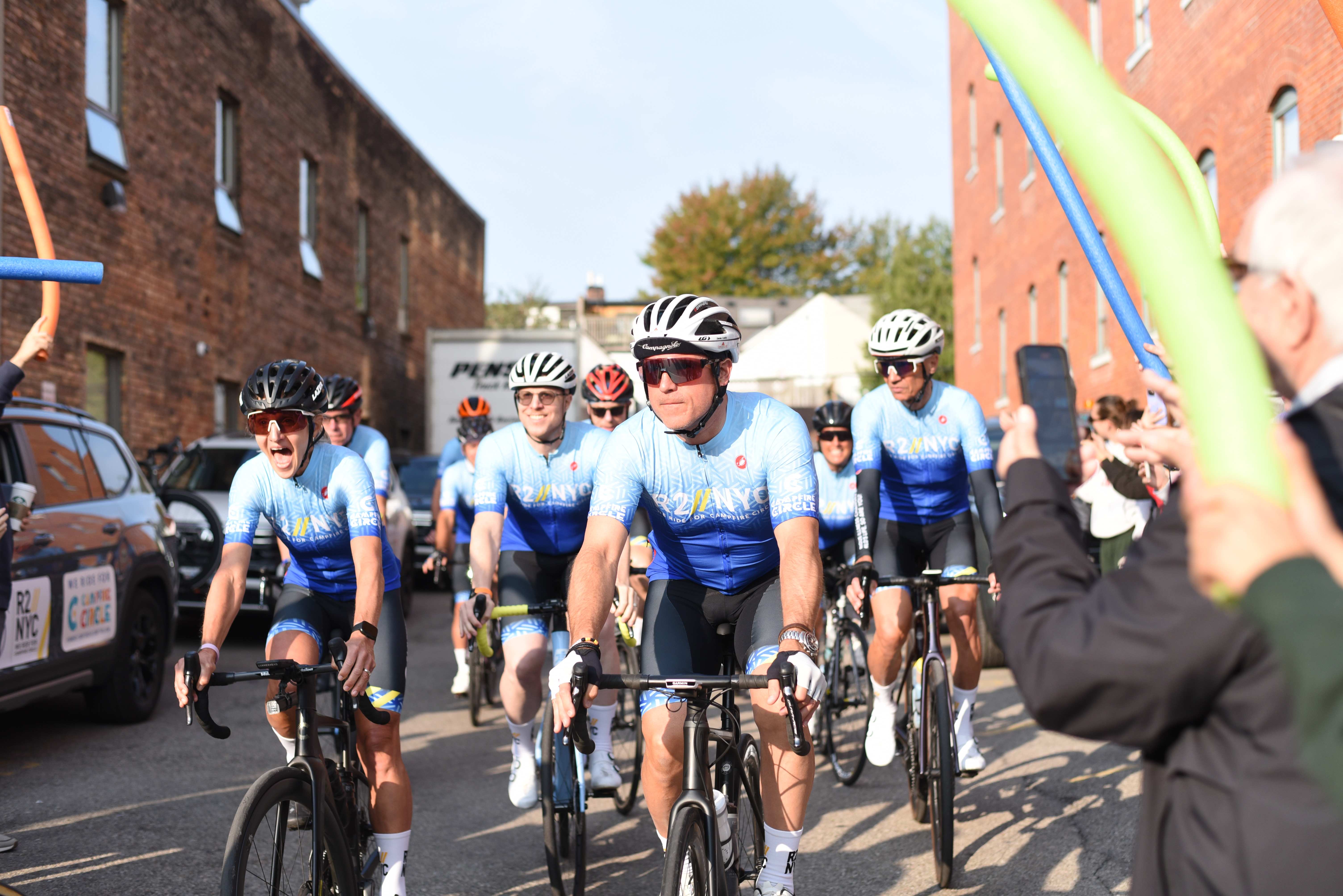 cyclists preparing to leave from parking lot with supporters cheering and holding pool noodles