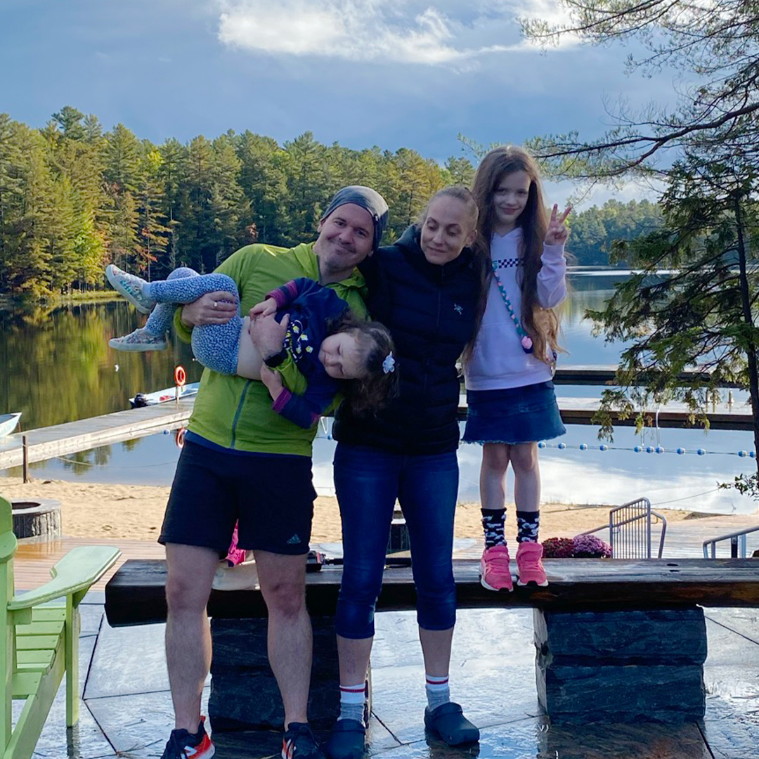 family standing together by the water at camp in muskoka with trees and lake in background