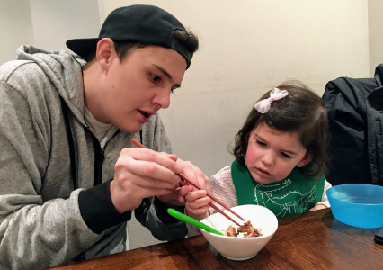 A young man named Andrew is showing a young girl how to hold chopsticks while sitting at a dining table.