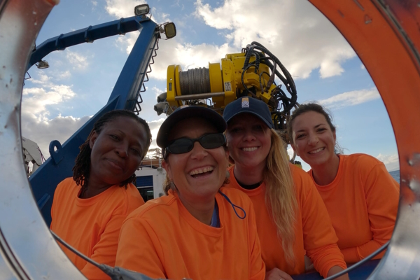Four women wearing orange smiling at the camera from a large, round metal hole. There is large yellow machinery behind them and the ocean.