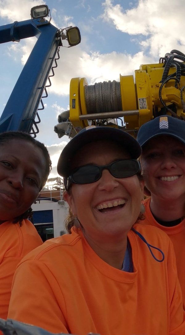 Three women wearing orange smiling at the camera from a large, round metal hole. There is large yellow machinery behind them and the ocean.