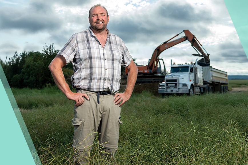 STARS Very Important Patient Sean Rickards stands on a worksite where the excavator and dump truck that pinned him work in the background.