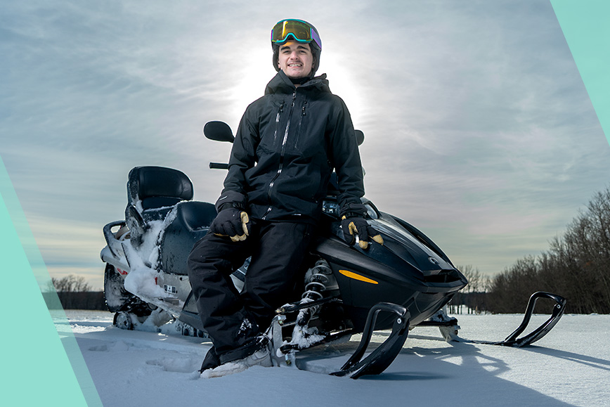 A teenage boy wearing a helmet and cold-weather gear leans against a stationary snowmobile.