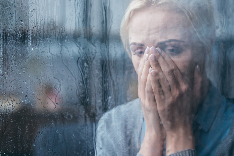 A women is upset and holds her hands to her mouth and looks out of a rainy window